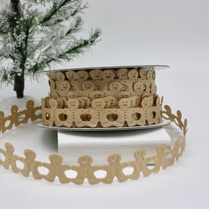 May include: A roll of tan gingerbread man ribbon sits on a white surface, with a garland of the same ribbon in front. A small, snow-covered Christmas tree is in the background. The ribbon features cut-out gingerbread men.
