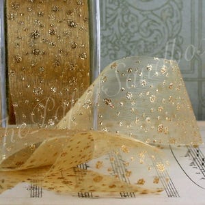 May include: A spool of gold ribbon with a sheer, dotted design. The ribbon is displayed with a portion unrolled, revealing its texture and the metallic gold accents. The ribbon is likely used for crafts, gift wrapping, or decorative purposes.