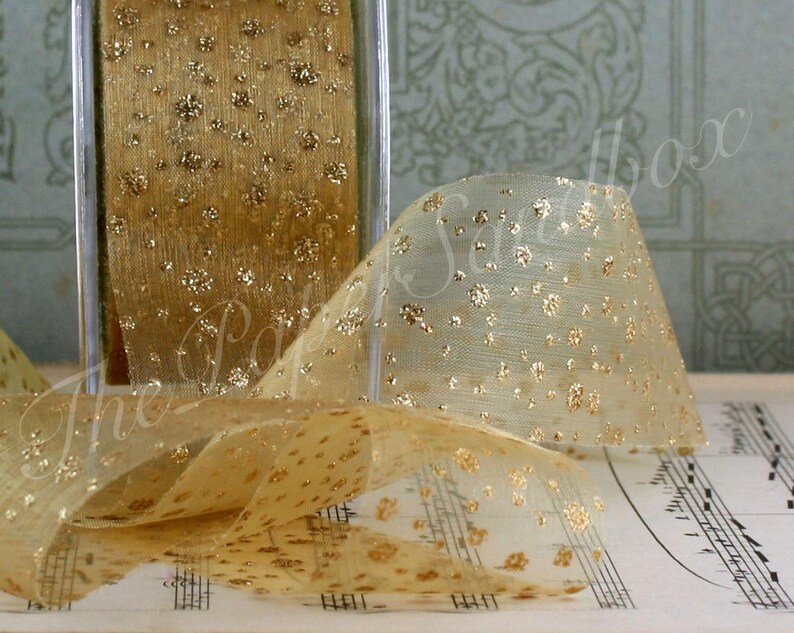 May include: A spool of gold ribbon with a sheer, dotted design. The ribbon is displayed with a portion unrolled, revealing its texture and the metallic gold accents. The ribbon is likely used for crafts, gift wrapping, or decorative purposes.