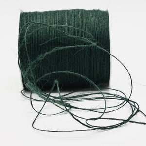 May include: A spool of dark green twine sits on a white surface. The twine is loosely wound, with strands extending from the spool. The color is a deep, forest green, suggesting a natural fiber product, suitable for crafts or packaging.