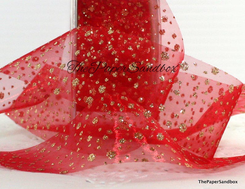 May include: Red sheer ribbon with gold glitter detailing. The ribbon is loosely arranged, showing a portion of the roll. Gold glitter is scattered across the ribbon, creating a festive appearance. The text "The Paper Sandbox" is visible.