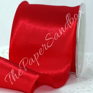 May include: A spool of vibrant red satin ribbon. The ribbon is glossy and appears to be unwound, revealing the shiny texture. The text "The PaperSandbox" is written across the ribbon in a cursive font.