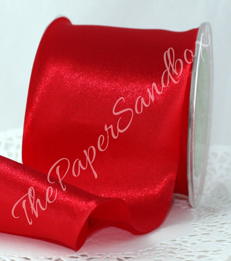 May include: A spool of vibrant red satin ribbon. The ribbon is glossy and appears to be unwound, revealing the shiny texture. The text "The PaperSandbox" is written across the ribbon in a cursive font.