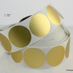 May include: A roll of gold metallic circle stickers, each 1.75 inches in diameter. The stickers are on a white backing and are partially unrolled, revealing the shiny gold surface. ThePaperSandbox is printed on the bottom right.