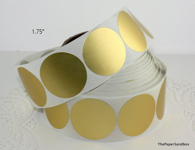 May include: A roll of gold metallic circle stickers, each 1.75 inches in diameter. The stickers are on a white backing and are partially unrolled, revealing the shiny gold surface. ThePaperSandbox is printed on the bottom right.