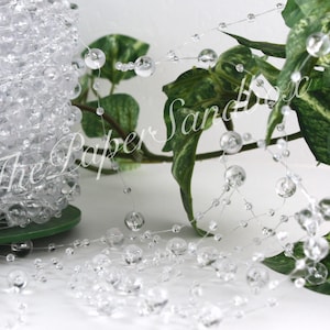 May include: A roll of clear, round, acrylic beads on a green spool. Strands of the beads are draped across a white surface, with green foliage in the background. The beads are various sizes and appear to be strung on clear wire.