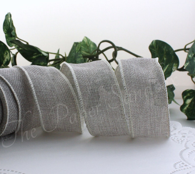 May include: A roll of gray burlap ribbon with a white edge, displayed on a white surface. The ribbon is loosely coiled, showcasing its texture and weave. Green leaves are visible in the background.