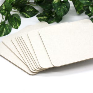 May include: A stack of square, off-white coasters with rounded corners. The coasters are made of a textured, absorbent material. Green leafy vines are in the background, adding a natural element to the image. The coasters are stacked in a fan shape.
