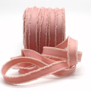 May include: A roll of pink ribbon with a textured, fringed edge. The ribbon is a solid pastel pink colour with a lighter, fringed trim. A length of the ribbon is unrolled and lies in front of the roll. The ribbon is likely used for crafts, gift wrapping, or decorative purposes.