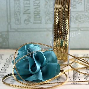 May include: A spool of gold sequin trim and a teal fabric flower are displayed on sheet music. The trim is in a clear plastic container. The Paper Sandbox is written on the image.