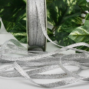 May include: A spool of silver ribbon with a woven texture. The ribbon is unwound, with some of it lying in loose loops. The ribbon is a light, metallic silver color, and is likely used for crafts or gift wrapping.