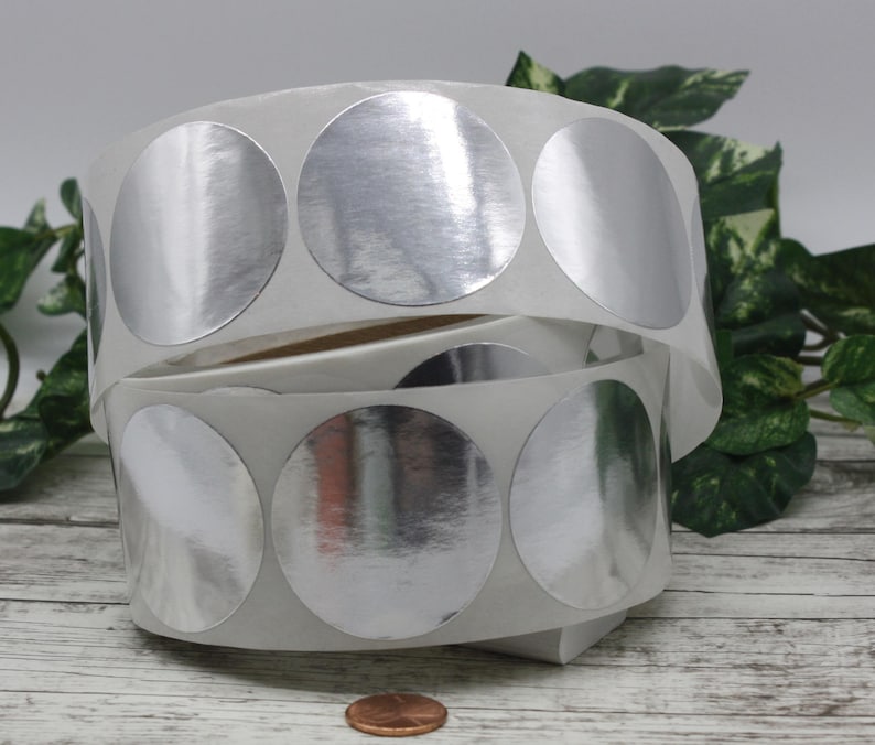May include: Two rolls of silver metallic circle stickers. The stickers are on a white backing and are partially unrolled, revealing the shiny surface. A penny is at the bottom of the image, providing a size reference. Green foliage is in the background.