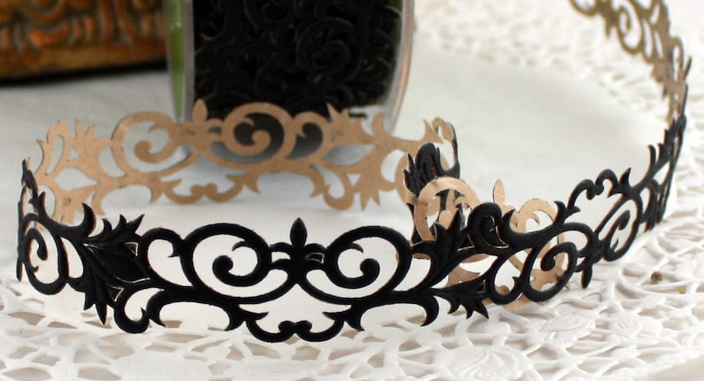 May include: A decorative paper trim with an ornate, scrollwork design. The trim is black on one side and tan on the other, with a repeating pattern of swirling lines and floral elements. The trim is likely used for scrapbooking or other crafts.