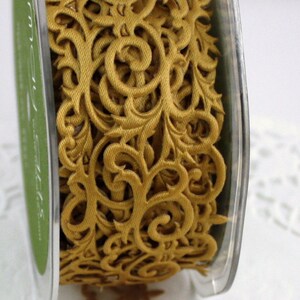 May include: A spool of gold-colored decorative ribbon with an intricate, openwork design. The ribbon features a repeating pattern of swirling floral and scroll motifs. The spool is encased in a clear plastic container.