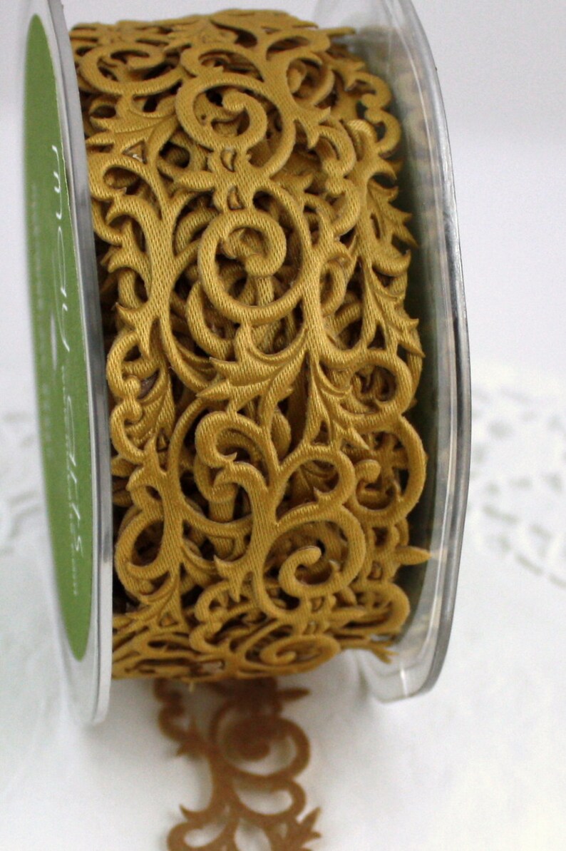 May include: A spool of gold-colored decorative ribbon with an intricate, openwork design. The ribbon features a repeating pattern of swirling floral and scroll motifs. The spool is encased in a clear plastic container.