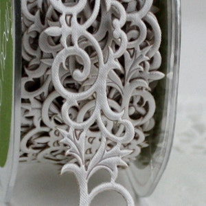 May include: A spool of white decorative ribbon with an intricate, openwork floral design. The ribbon features scrolling patterns and leaf-like shapes, perfect for embellishing crafts or gifts. The ribbon is made of a soft, fabric-like material.