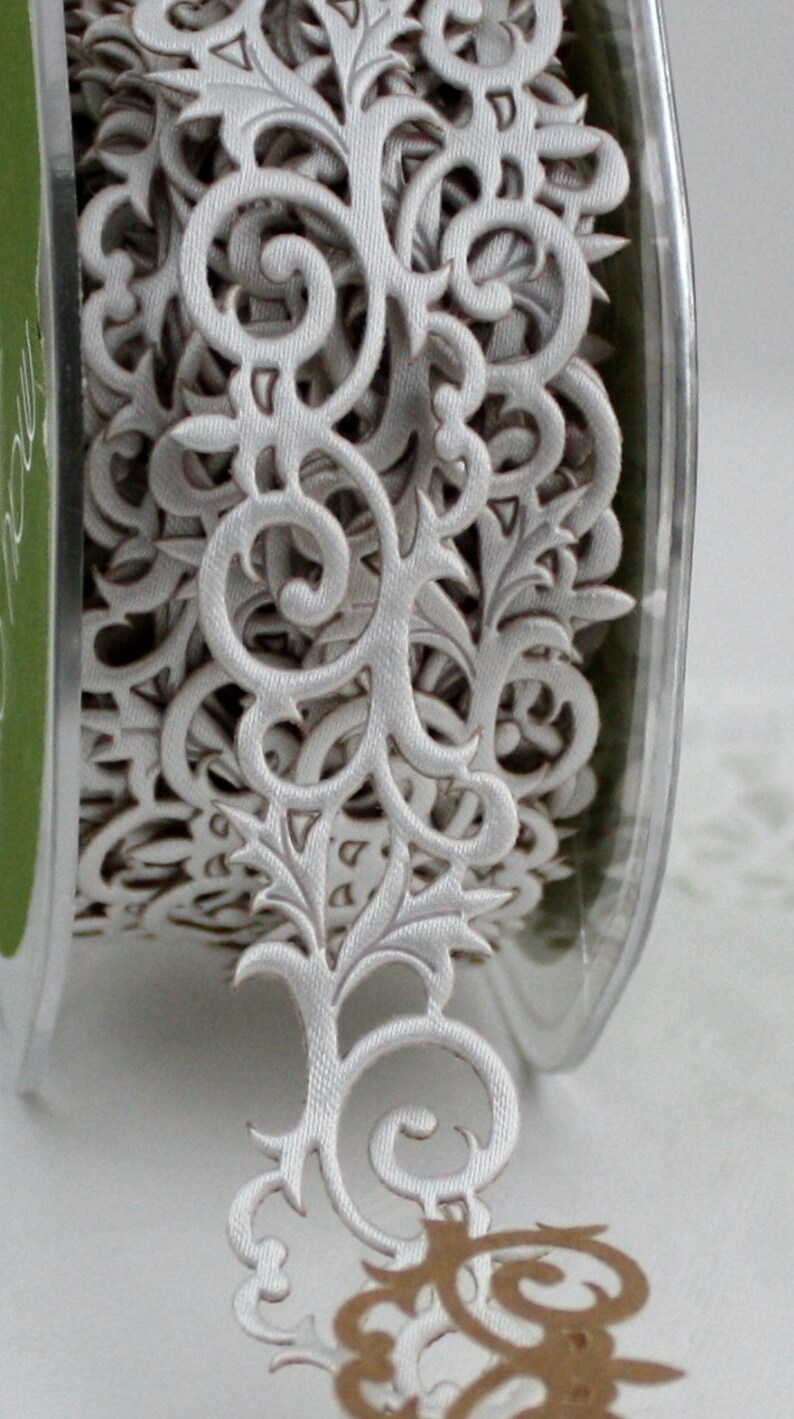May include: A spool of white decorative ribbon with an intricate, openwork floral design. The ribbon features scrolling patterns and leaf-like shapes, perfect for embellishing crafts or gifts. The ribbon is made of a soft, fabric-like material.