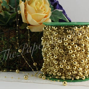 May include: A spool of gold beaded garland with a green base. The garland is made of small and large gold beads strung together. The garland is next to a floral arrangement with yellow roses.