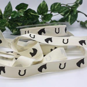 May include: Cream-colored ribbon with a repeating pattern of black horseshoes and horse heads. The ribbon is unspooled, with a roll of ribbon visible in the background. Green foliage is in the background.