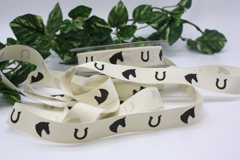 May include: Cream-colored ribbon with a repeating pattern of black horseshoes and horse heads. The ribbon is unspooled, with a roll of ribbon visible in the background. Green foliage is in the background.