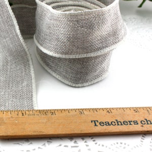 May include: A roll of gray burlap ribbon with white edging, next to a wooden ruler. The ruler reads "Teachers change lives". The ribbon is approximately 2 inches wide.