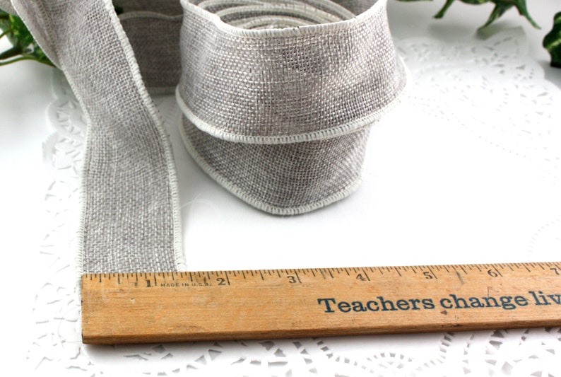 May include: A roll of gray burlap ribbon with white edging, next to a wooden ruler. The ruler reads "Teachers change lives". The ribbon is approximately 2 inches wide.