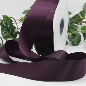 May include: A roll of deep purple satin ribbon. The ribbon is unwound, with a portion of the spool visible. Green leafy vines are in the background. The ribbon has a smooth, shiny texture.