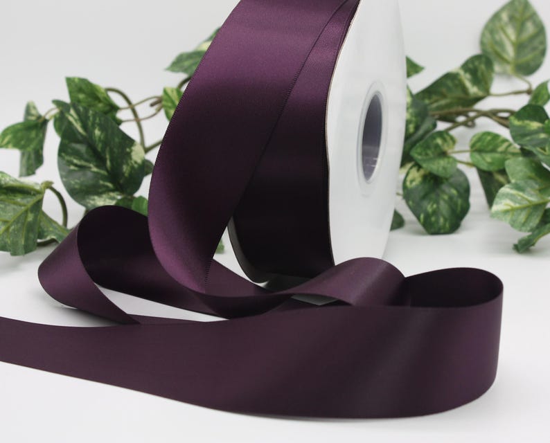 May include: A roll of deep purple satin ribbon. The ribbon is unwound, with a portion of the spool visible. Green leafy vines are in the background. The ribbon has a smooth, shiny texture.