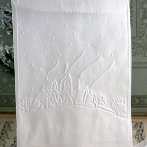 Op de afbeelding: Witte papieren cadeauzak met een reli&euml;f winterdorpssc&egrave;ne. Het ontwerp toont een rij huizen, bomen en vallende sneeuw. De bovenrand van de zak heeft een decoratieve, geschulpte rand.