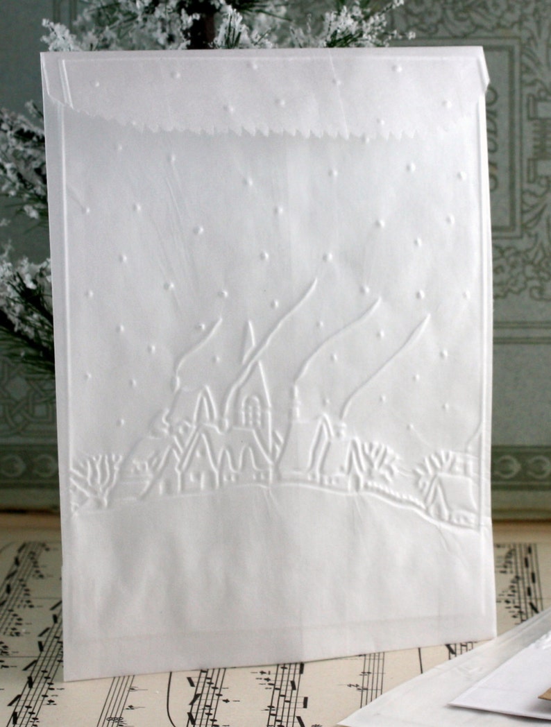Op de afbeelding: Witte papieren cadeauzak met een reli&euml;f winterdorpssc&egrave;ne. Het ontwerp toont een rij huizen, bomen en vallende sneeuw. De bovenrand van de zak heeft een decoratieve, geschulpte rand.