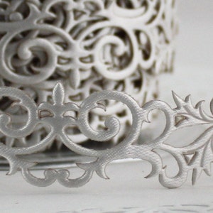May include: Close-up of a roll of silver decorative paper ribbon with an intricate, openwork floral design. The ribbon is partially unrolled, revealing a detailed pattern of swirling leaves and flourishes, perfect for crafts and embellishments.