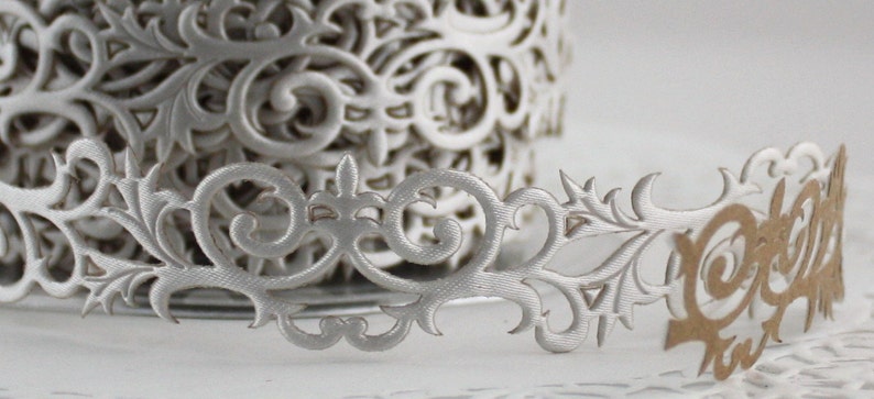 May include: Close-up of a roll of silver decorative paper ribbon with an intricate, openwork floral design. The ribbon is partially unrolled, revealing a detailed pattern of swirling leaves and flourishes, perfect for crafts and embellishments.