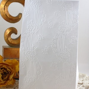 May include: A white card with an embossed floral and scroll design. The card has a rectangular shape and is standing upright. The design is repeated across the surface. A gold-colored decorative object and a dried yellow rose are in the background.