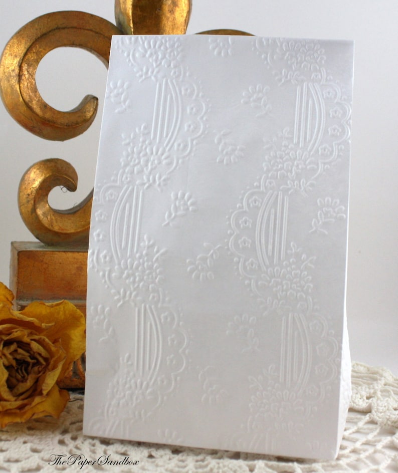 May include: A white card with an embossed floral and scroll design. The card has a rectangular shape and is standing upright. The design is repeated across the surface. A gold-colored decorative object and a dried yellow rose are in the background.