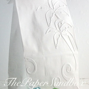 May include: A white paper gift bag with an embossed palm tree design. The bag has a folded top and is decorated with a stylized palm tree and swirling accents. The text "The Paper Sandbox" is visible.