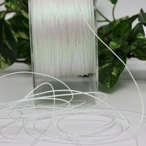 May include: A spool of white, iridescent thread. The thread is tightly wound on a clear plastic spool. Strands of the thread are unwound and scattered in the foreground. The thread has a subtle, shimmering quality, reflecting light.