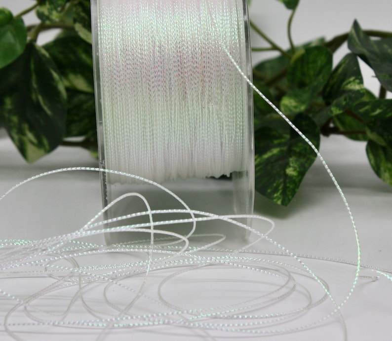 May include: A spool of white, iridescent thread. The thread is tightly wound on a clear plastic spool. Strands of the thread are unwound and scattered in the foreground. The thread has a subtle, shimmering quality, reflecting light.