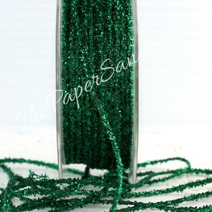 May include: A spool of emerald green glitter ribbon. The ribbon is tightly wound on a clear plastic spool, with some of the ribbon unwound and lying in front. The glitter catches the light, creating a sparkling effect. The Paper Sandbox is written on the image.