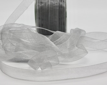May include: A spool of silver-gray sheer ribbon is unraveled, with the ribbon cascading across a white surface. The ribbon has a delicate, translucent quality, and a roll of the ribbon is visible in the background.