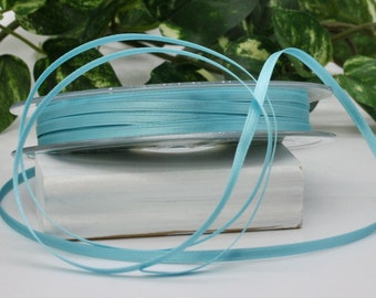 May include: Spools of light blue ribbon. The ribbon is thin and appears to be made of satin. The spools are stacked on top of a white rectangular object. Green foliage is in the background.