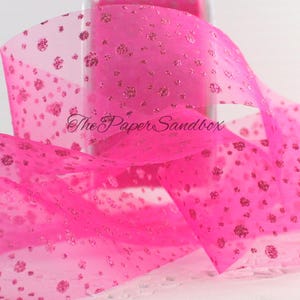 May include: Bright pink sheer ribbon with glitter accents. The ribbon has a delicate, dotted pattern and the text "The Paper Sandbox" is visible. This decorative ribbon is suitable for crafts, gift wrapping, and event decorations.