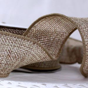 May include: Close-up of a roll of natural-colored burlap ribbon. The ribbon has a woven texture with a brown edge. The ribbon is curled, showing the front and back of the material. This ribbon is suitable for crafts and decorations.