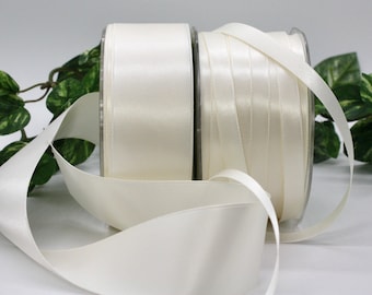 May include: Two spools of ivory-colored ribbon are displayed, one with a satin finish and the other with a ribbed texture. The ribbon is unrolled, showing the smooth, shiny surface. Green foliage is visible in the background.