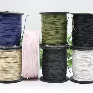 May include: An assortment of colorful spools of cord, including navy blue, tan, pink, olive green, black, and white. The spools are stacked, showcasing the variety of colors and textures available for crafting and DIY projects.