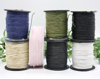 May include: An assortment of colorful spools of cord, including navy blue, tan, pink, olive green, black, and white. The spools are stacked, showcasing the variety of colors and textures available for crafting and DIY projects.