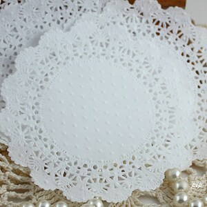 May include: Close-up of several white paper doilies. The doilies have a scalloped edge with a delicate, lacy design. The center of each doily has a subtle dotted texture. The doilies are stacked on top of each other.