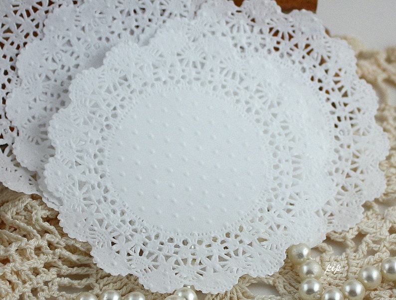 May include: Close-up of several white paper doilies. The doilies have a scalloped edge with a delicate, lacy design. The center of each doily has a subtle dotted texture. The doilies are stacked on top of each other.