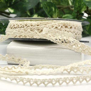 May include: A spool of off-white lace trim featuring a repeating zigzag or rickrack pattern along the edges. The delicate, woven trim is wound around a clear plastic reel, with a length of the lace extending forward in a loose, wavy line.