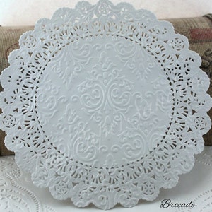 May include: A white paper doily with a detailed, embossed floral design in the center and a scalloped, lace-like edge. The word "Brocade" is printed in the bottom right corner.