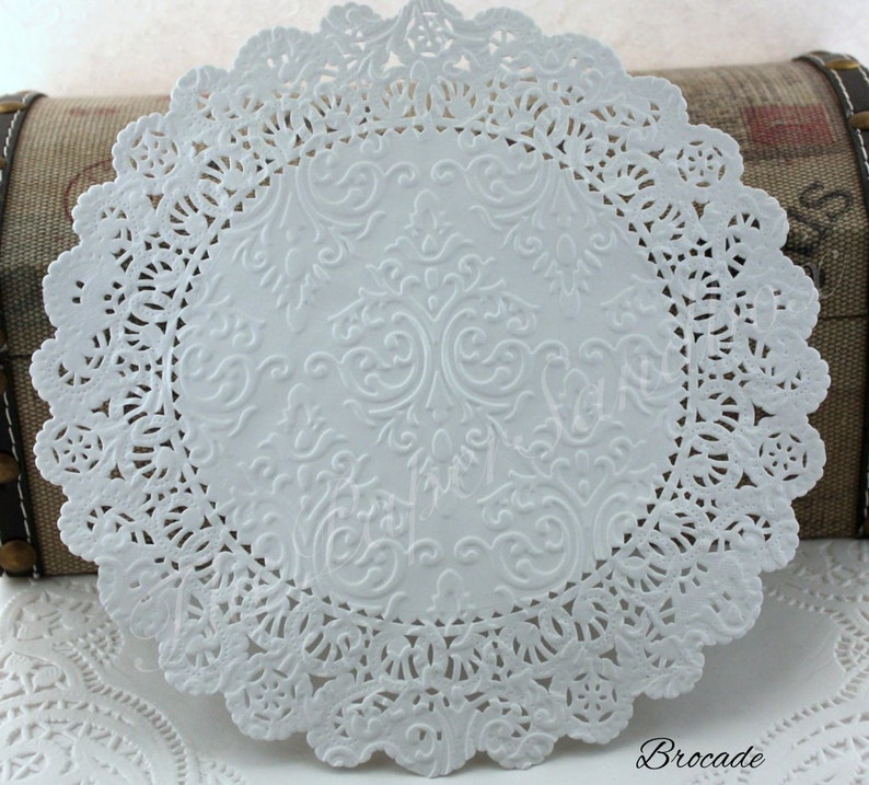 May include: A white paper doily with a detailed, embossed floral design in the center and a scalloped, lace-like edge. The word "Brocade" is printed in the bottom right corner.